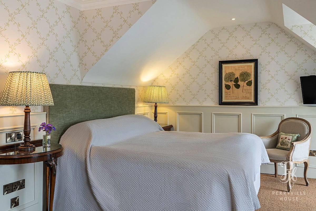 A neatly arranged bedroom with a large bed, two bedside lamps, a framed botanical print on the wall, and a chair with a cushion.