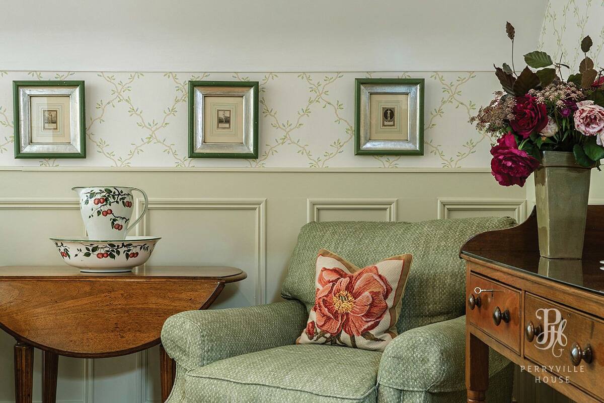 A room with a green upholstered armchair, a wooden side table with a floral-patterned bowl and cup, and a vase with flowers on a dresser.