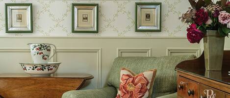 A room with a green upholstered armchair, a wooden side table with a floral-patterned bowl and cup, and a vase with flowers on a dresser.