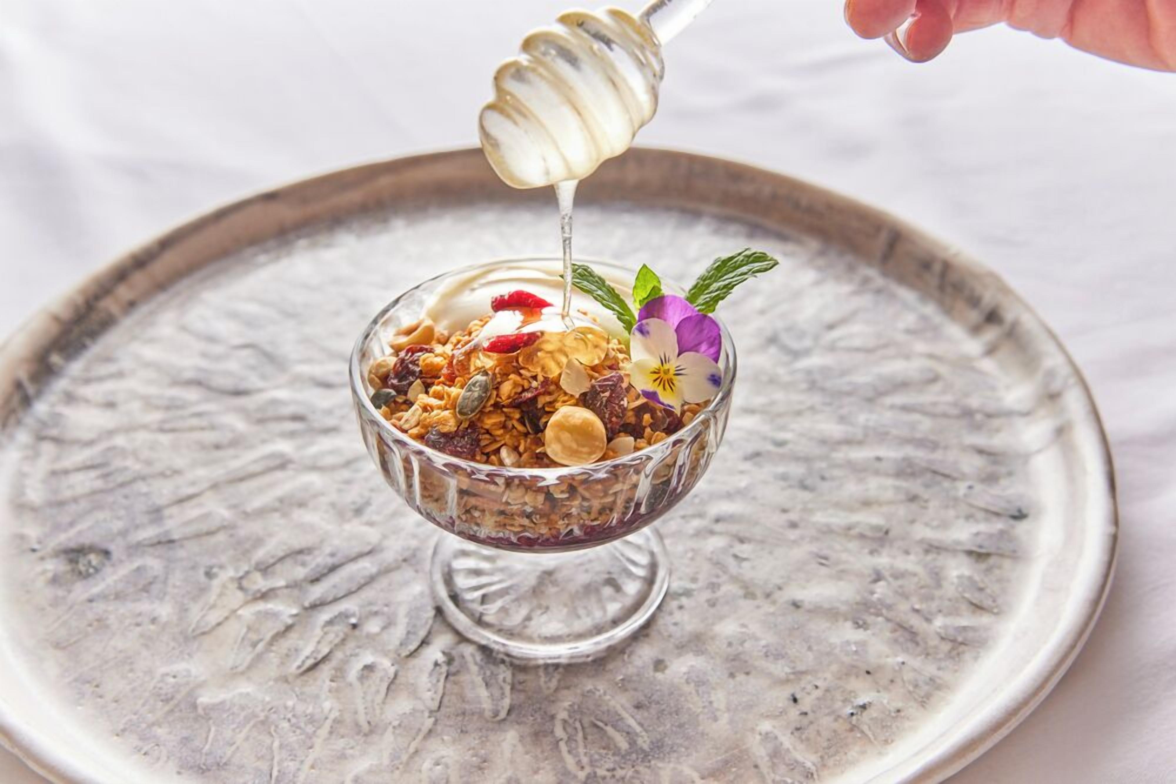 A bowl of granola with honey being drizzled over it, placed on a decorative plate.
