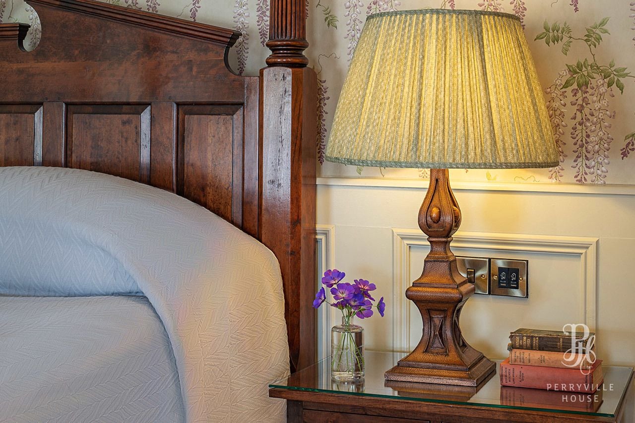Een net opgemaakt bed met houten hoofdbord, een glazen bijzettafeltje met een lamp, een vaas met paarse bloemen en een stapel boeken.