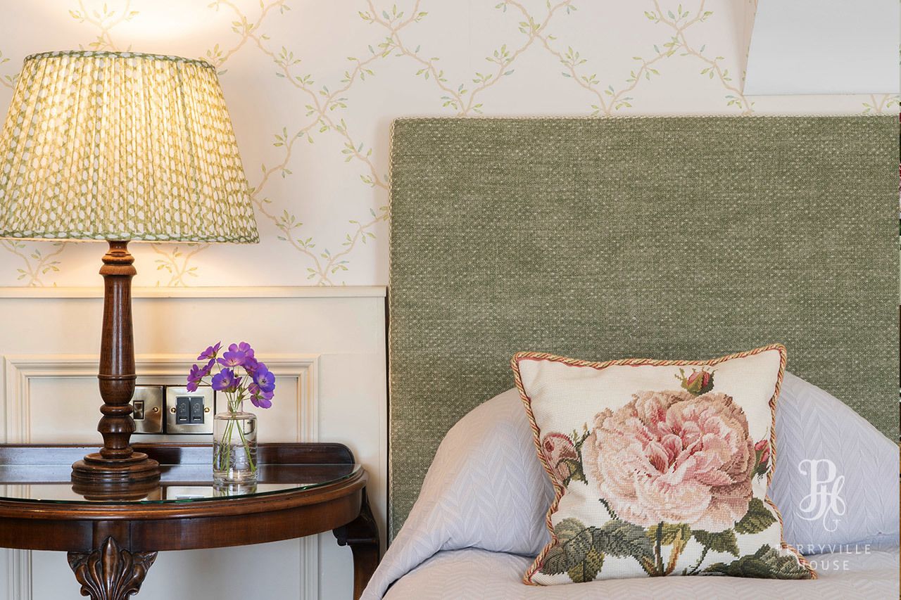 A neatly arranged bedroom with a green headboard, a bedside table with a lamp and vase of flowers, and a floral pillow.