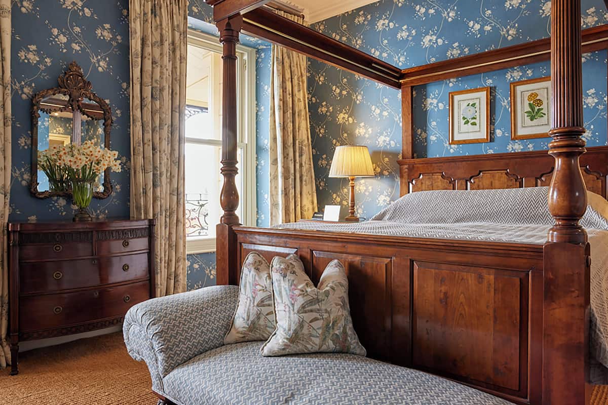 A four-poster bed with floral-patterned pillows, a wooden dresser, and a mirror with a vase of flowers.