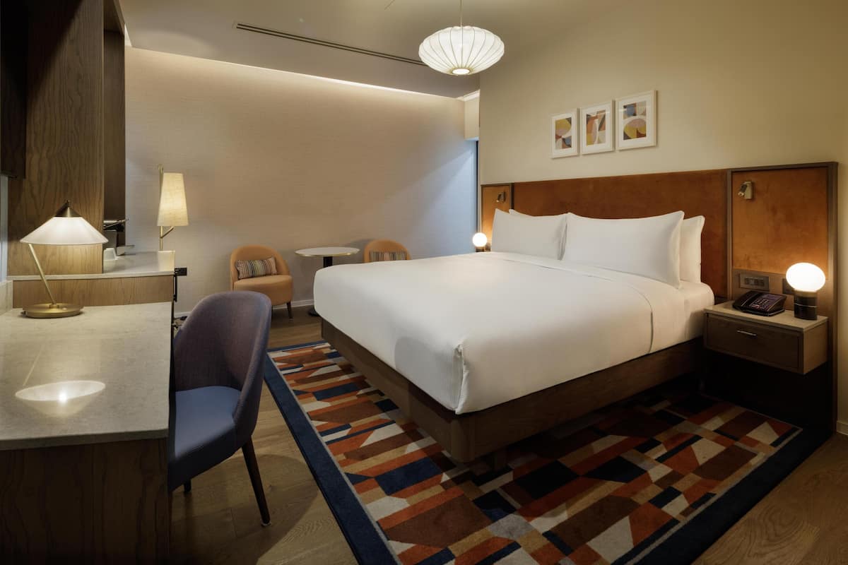 A hotel room with a large bed, a desk with a lamp, a chair, a bedside table with a phone, and a patterned rug.