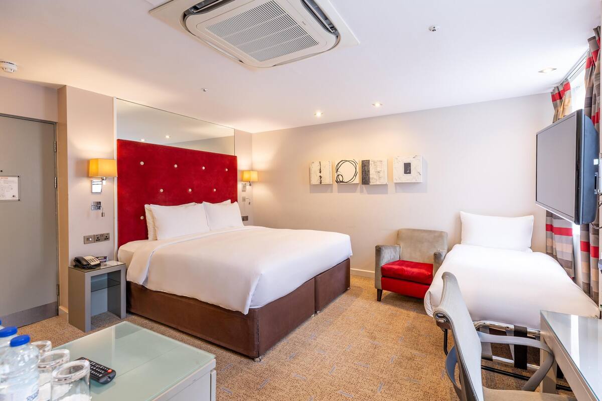 A hotel room with a bed, a desk, a chair, a mirror, a red accent wall, and a television.