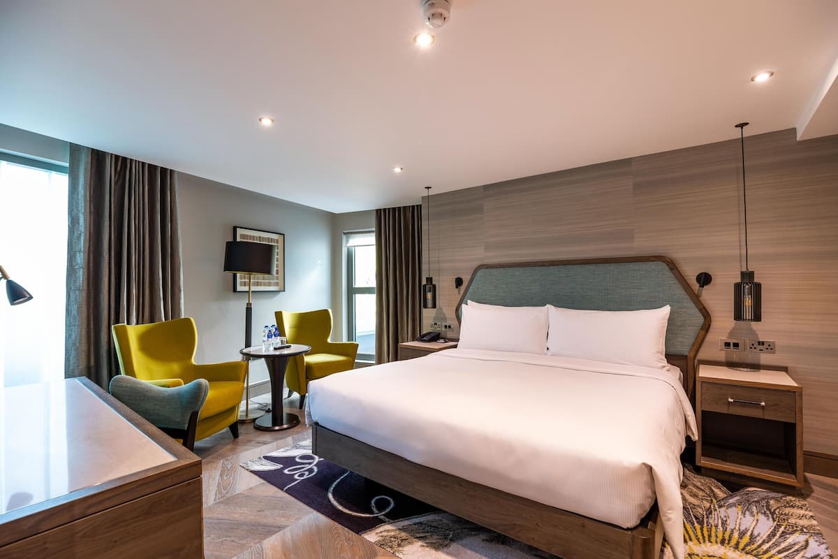 A modern hotel room with a large bed, a desk, two chairs, a small table, and a nightstand with a lamp.