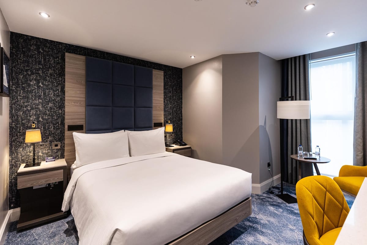 A hotel room with a large bed, bedside tables, a lamp, a small table with a chair, and a view of the outdoors.