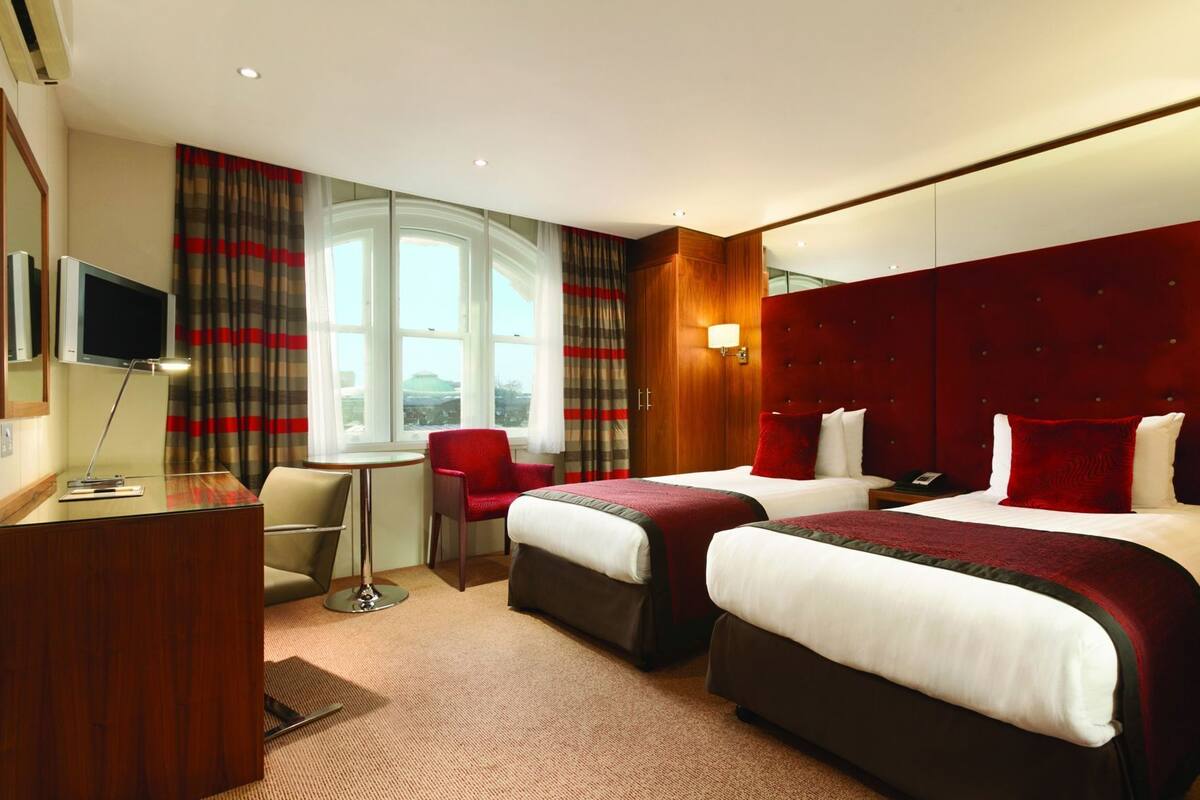 A hotel room with two beds, a desk, a chair, a TV, and a window with curtains.