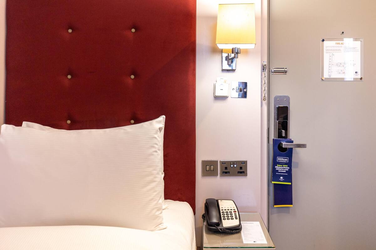 A hotel room with a tufted headboard, a bedside telephone, a lamp, a fire alarm, and a door with a keycard reader.