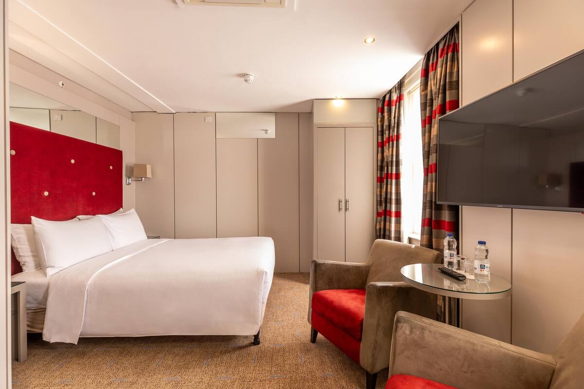 A hotel room with a bed, a TV, a small table with water bottles, a chair, and a wardrobe.