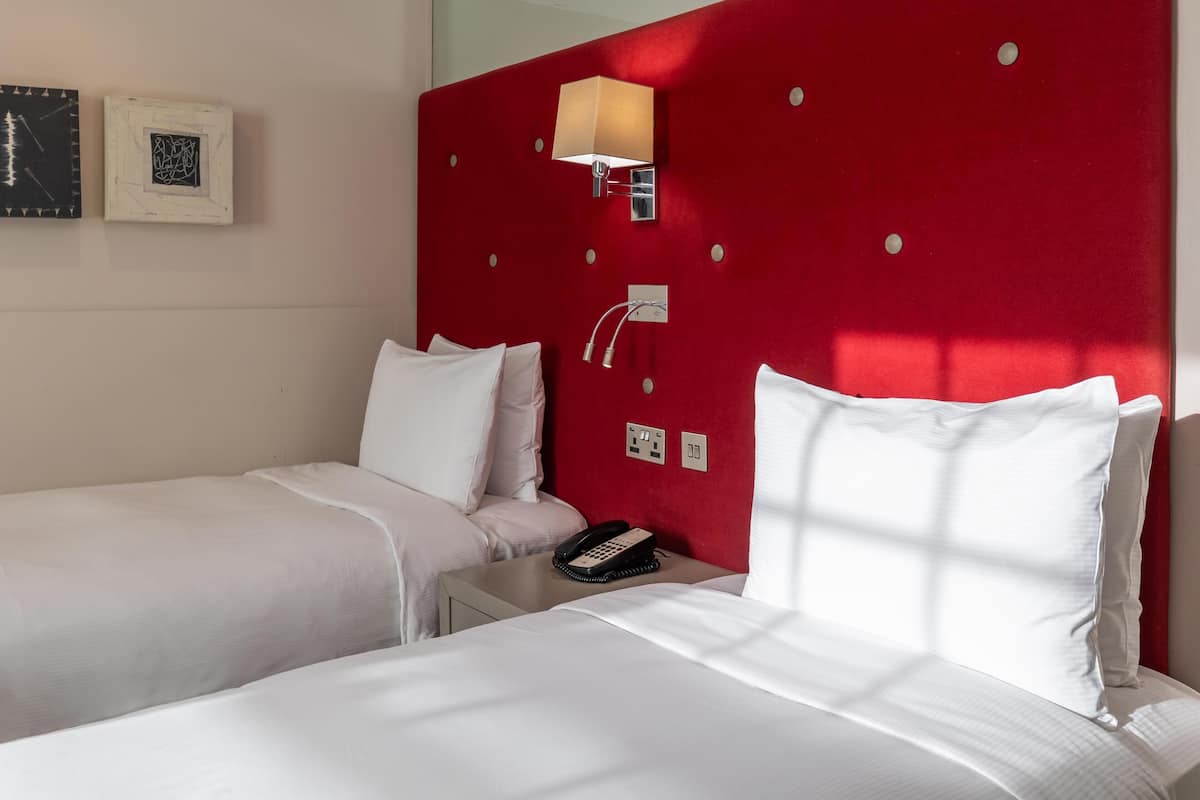 A hotel room with two beds, a red headboard, a bedside lamp, a phone, and wall art.