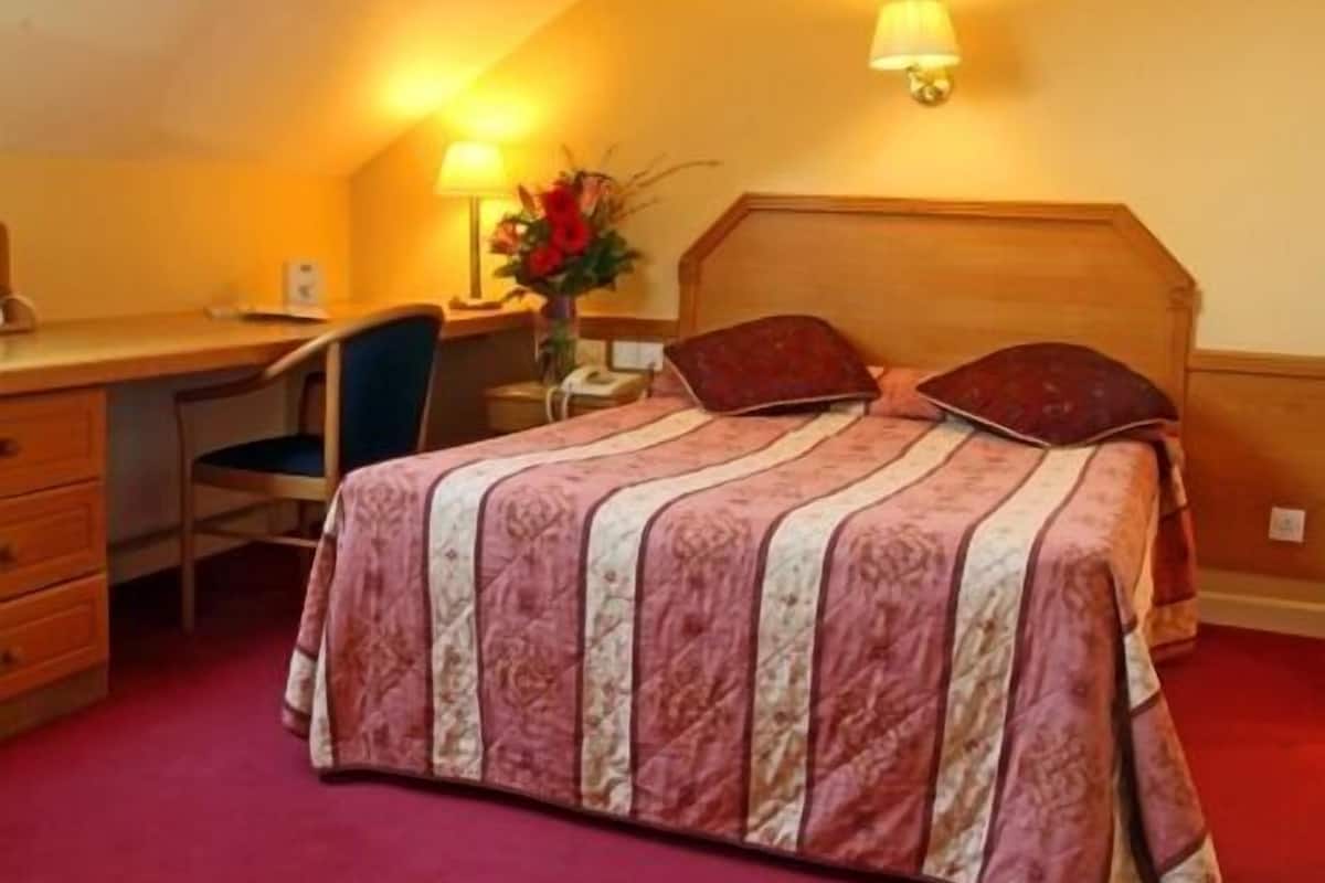 Un dormitorio con techo inclinado, una cama de madera con colcha estampada, un escritorio con silla y una lámpara de pared.