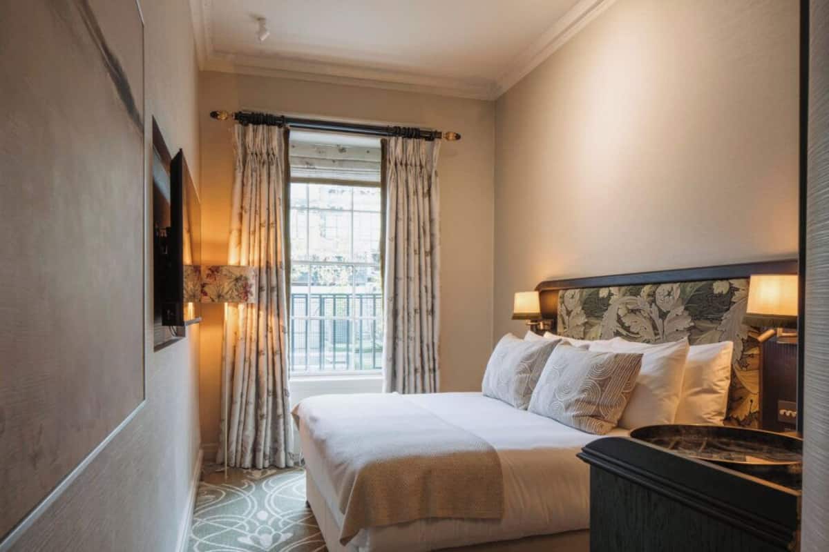 A hotel room with a bed, a nightstand, a lamp, a window with curtains, and a view of the outside.