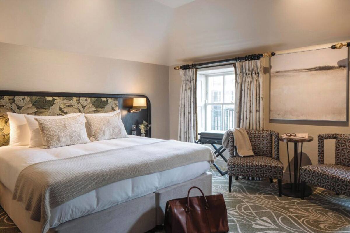 A hotel room with a large bed, a nightstand, a chair, a small table, and a window with curtains.