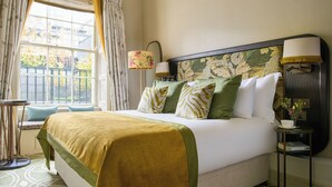 Una cama bien hecha con un cabecero verde y blanco estampado, una mesita de noche con una lámpara y una ventana con cortinas.