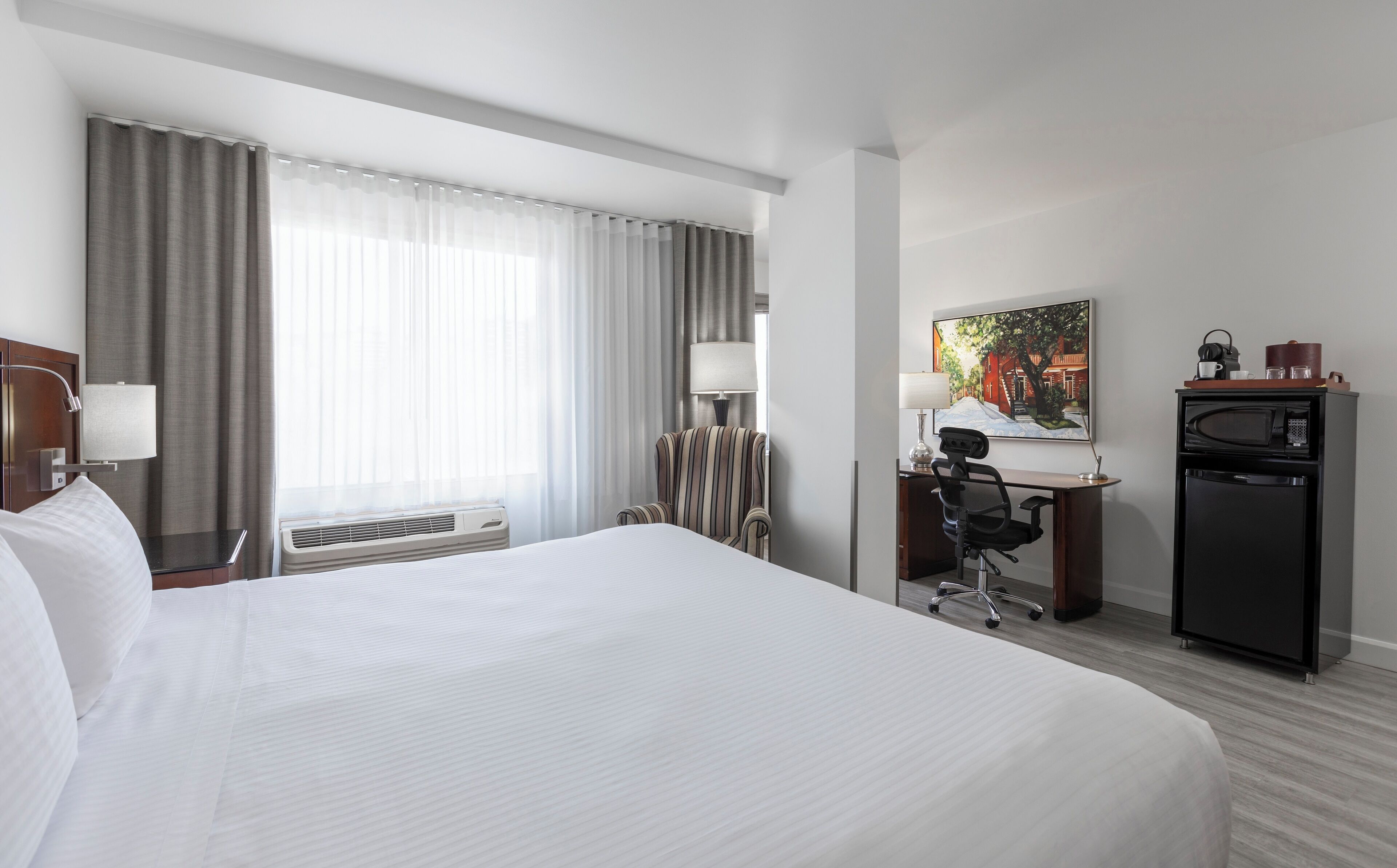 A hotel room with a large bed, a desk with a chair, a desk lamp, a refrigerator, and a painting on the wall.