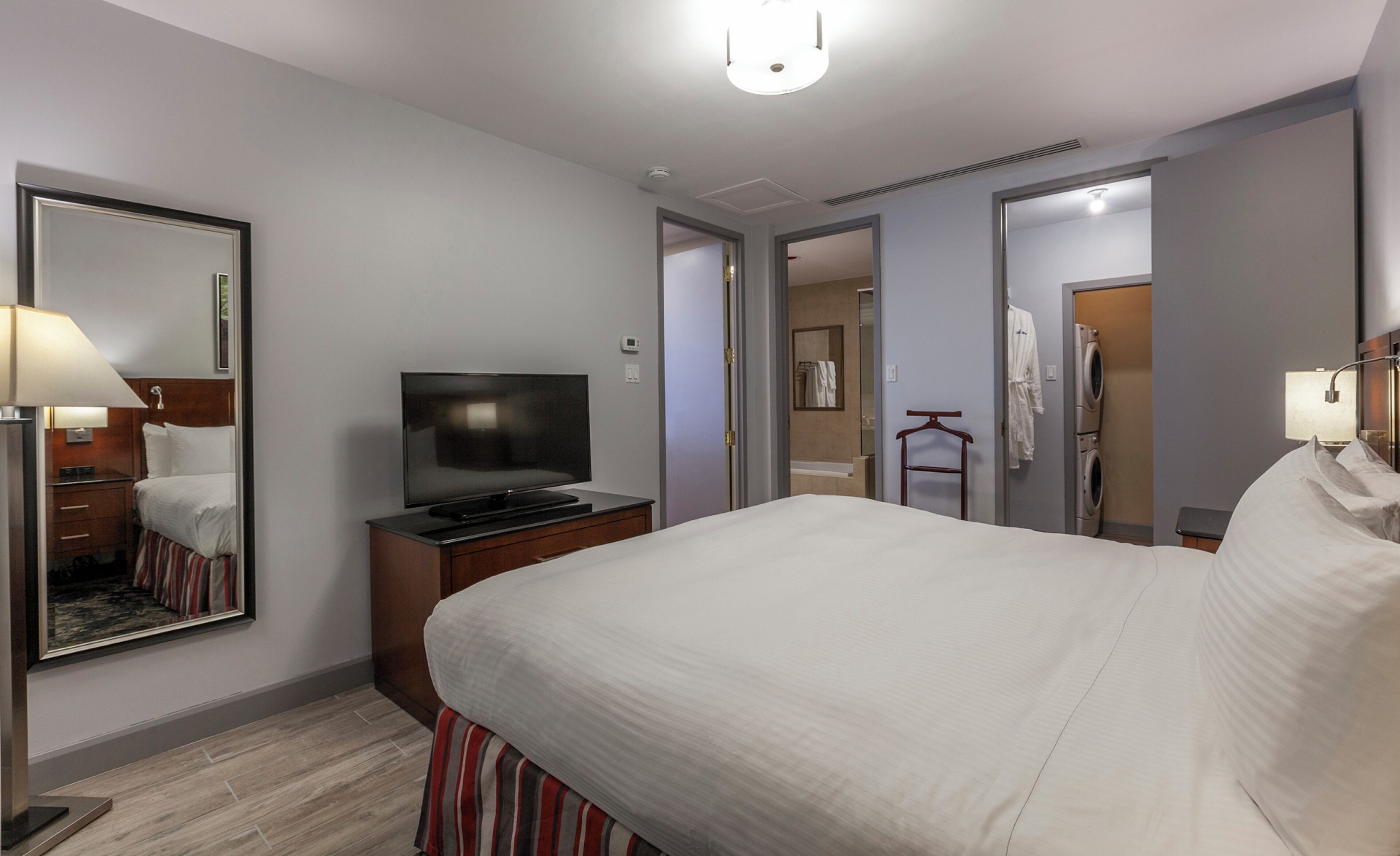 A hotel room with a large bed, a TV, a mirror, a dresser, and a bathroom visible through an open door.