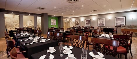 Comedor de hotel con varias mesas dispuestas para el desayuno, con platos blancos, tazas y cubiertos.