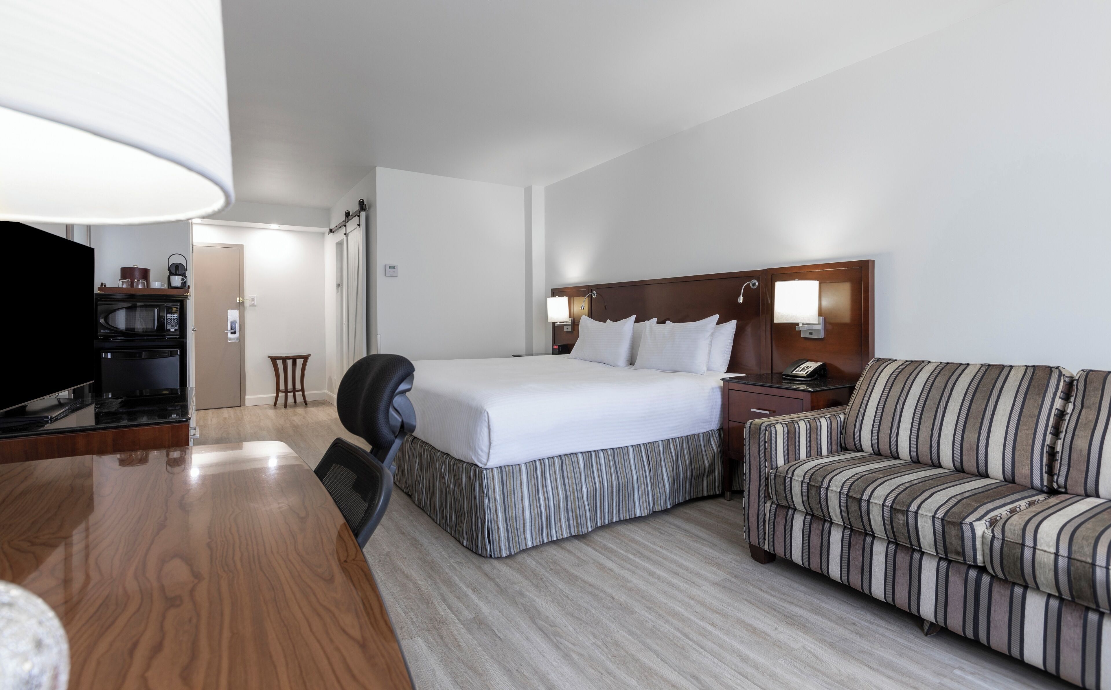 Habitación de hotel con cama, sofá, escritorio con lámpara, televisor y kitchenette con microondas y hervidor.