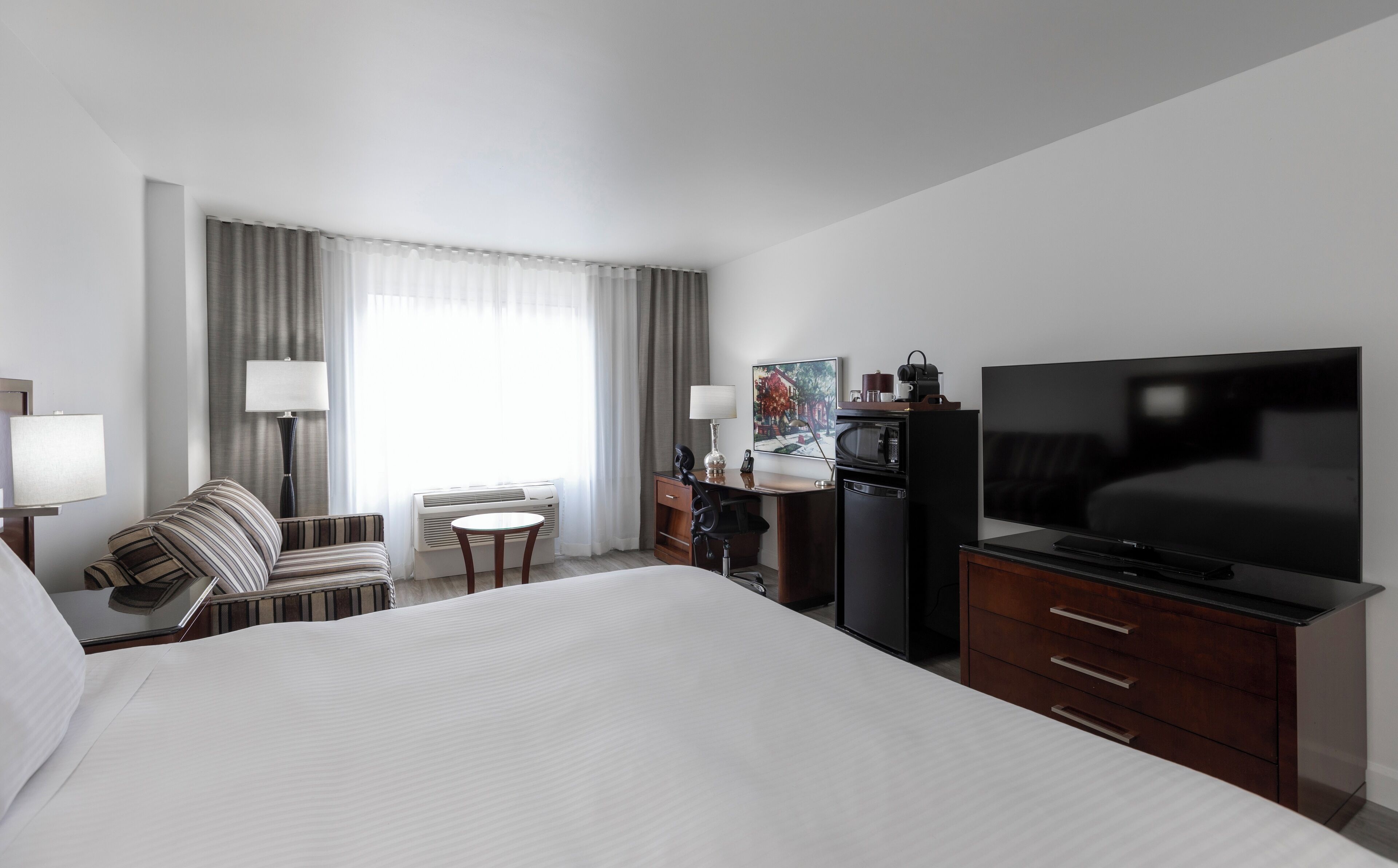 Habitación de hotel con una cama grande, un escritorio con televisor, una silla, una lámpara y una ventana con cortinas.