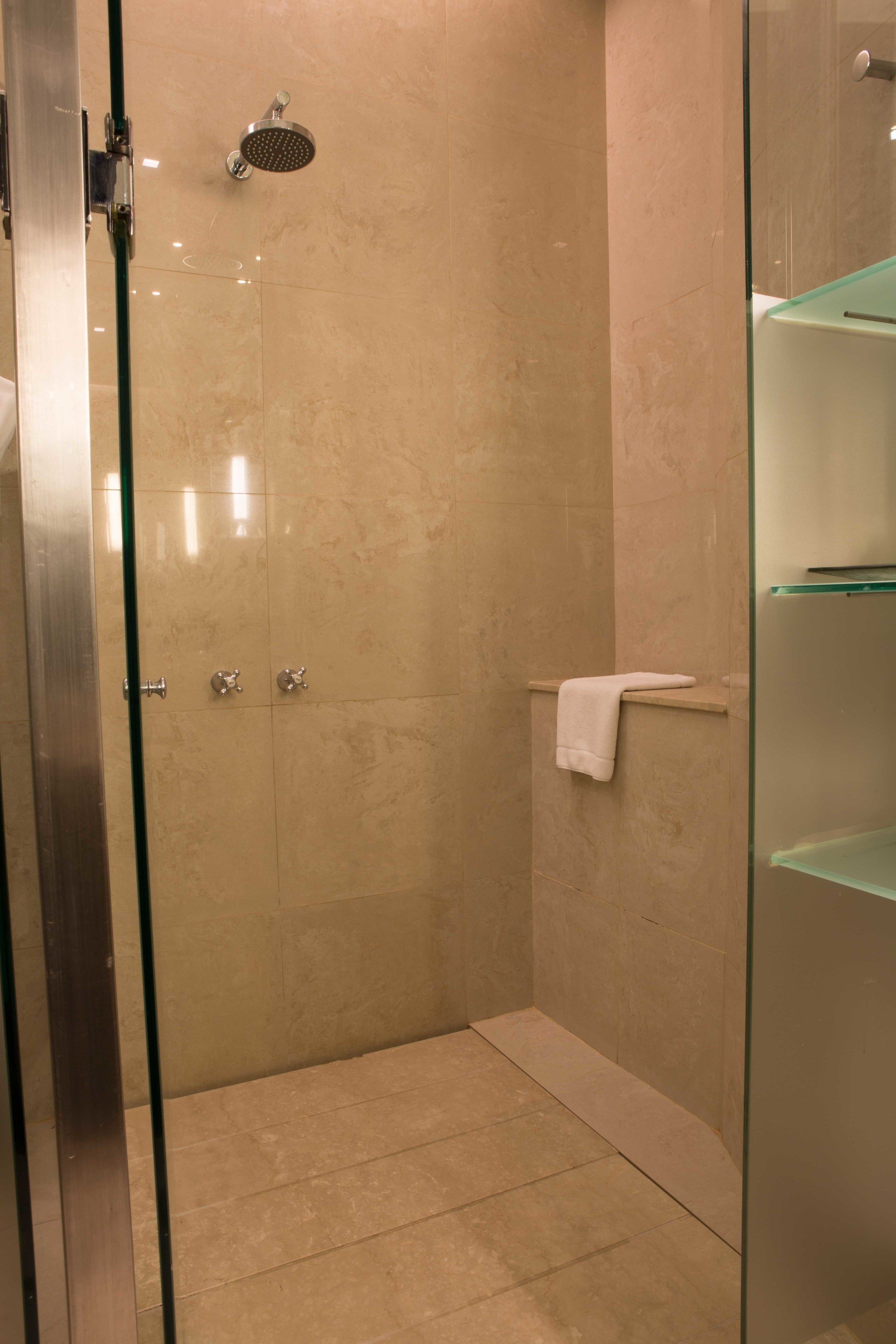 A modern bathroom with a glass-enclosed shower, beige tiles, and a glass shelf unit.
