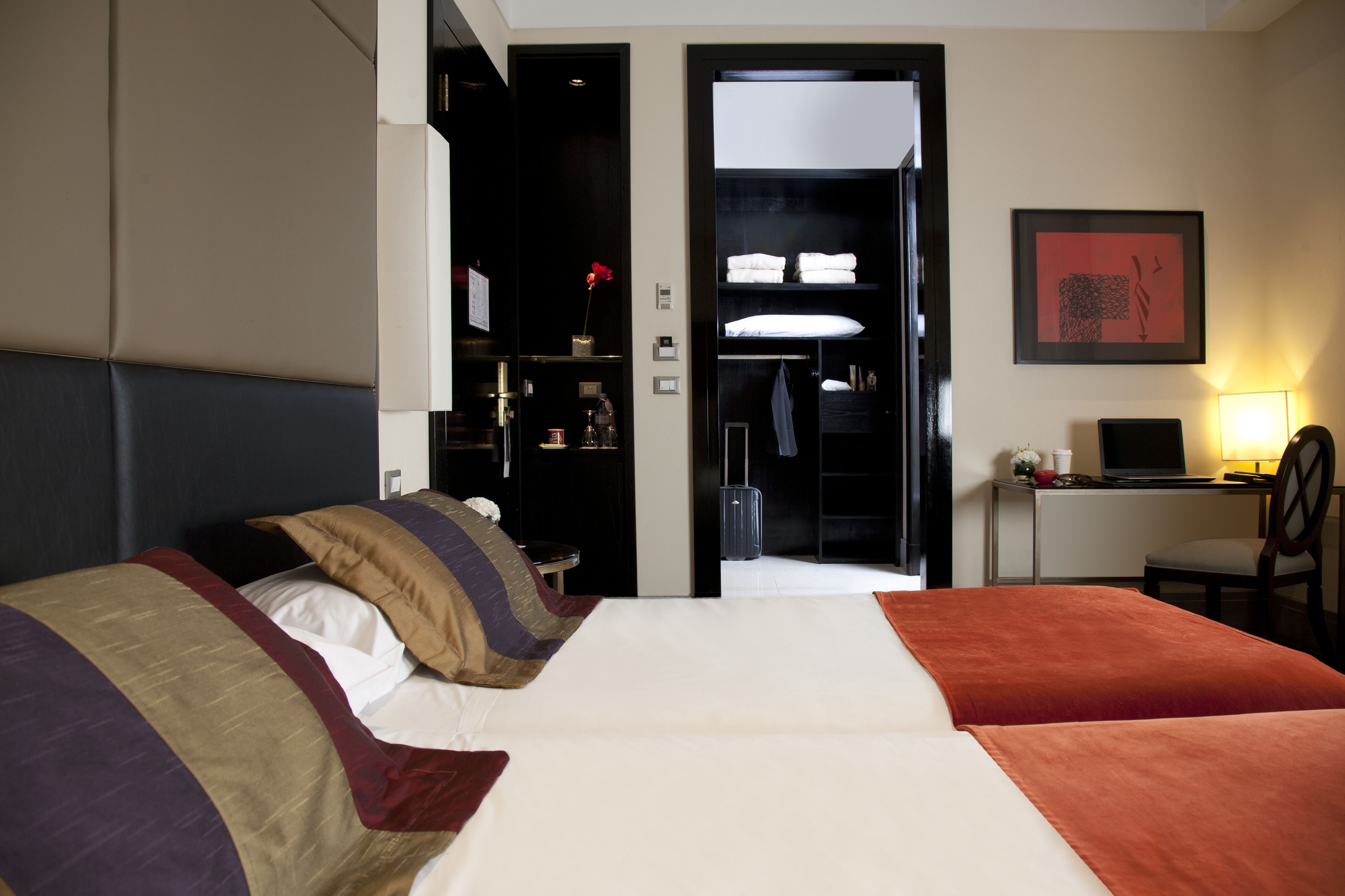Ein Hotelzimmer mit zwei Betten, einem Schreibtisch mit einem Laptop, einem Stuhl, einem Kleiderschrank und einem gerahmten Bild an der Wand.