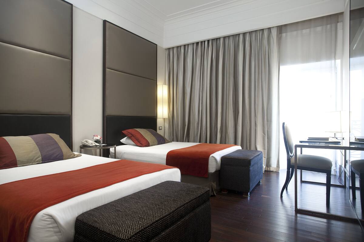 Habitación de hotel con dos camas, un escritorio, una silla y un amplio ventanal con cortinas.
