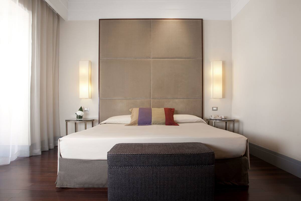 Una habitación de hotel moderna con una cama grande, dos mesitas de noche, un banco y un amplio ventanal con cortinas.