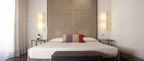 Una habitación de hotel moderna con una cama grande, dos mesitas de noche, un banco y un amplio ventanal con cortinas.