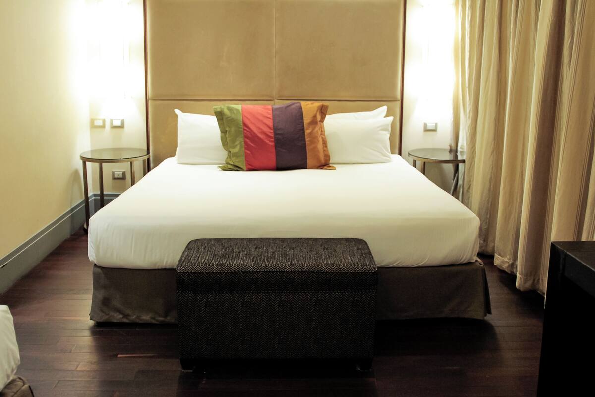 Habitación de hotel con una cama grande, dos mesitas de noche, un banco y una ventana con cortinas.
