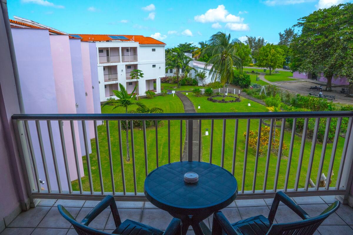 A balcony with a table and chairs, overlooking a well-maintained garden and a building with multiple balconies.