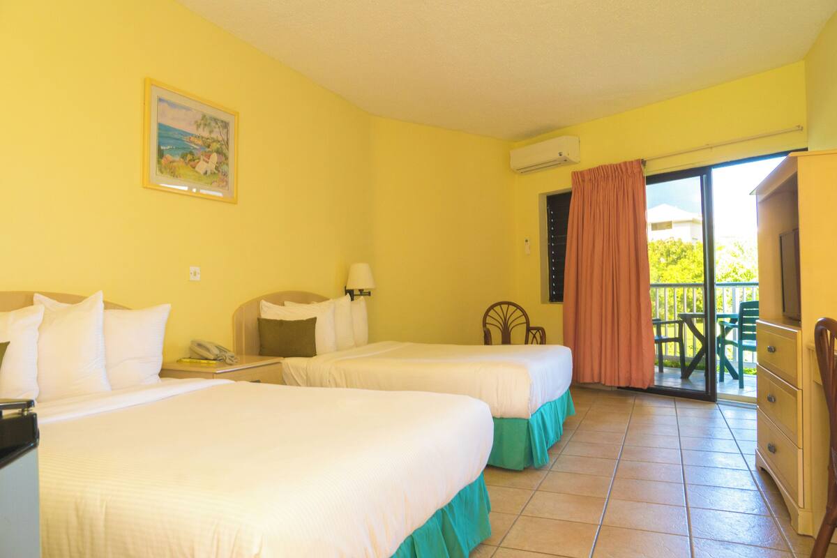 A hotel room with two beds, a desk, a chair, a television, and a balcony with a view of greenery.