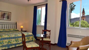 Habitación de hotel con una cama, una mesa y una silla. Se ve una ventana con cortinas azules y vistas a un edificio.