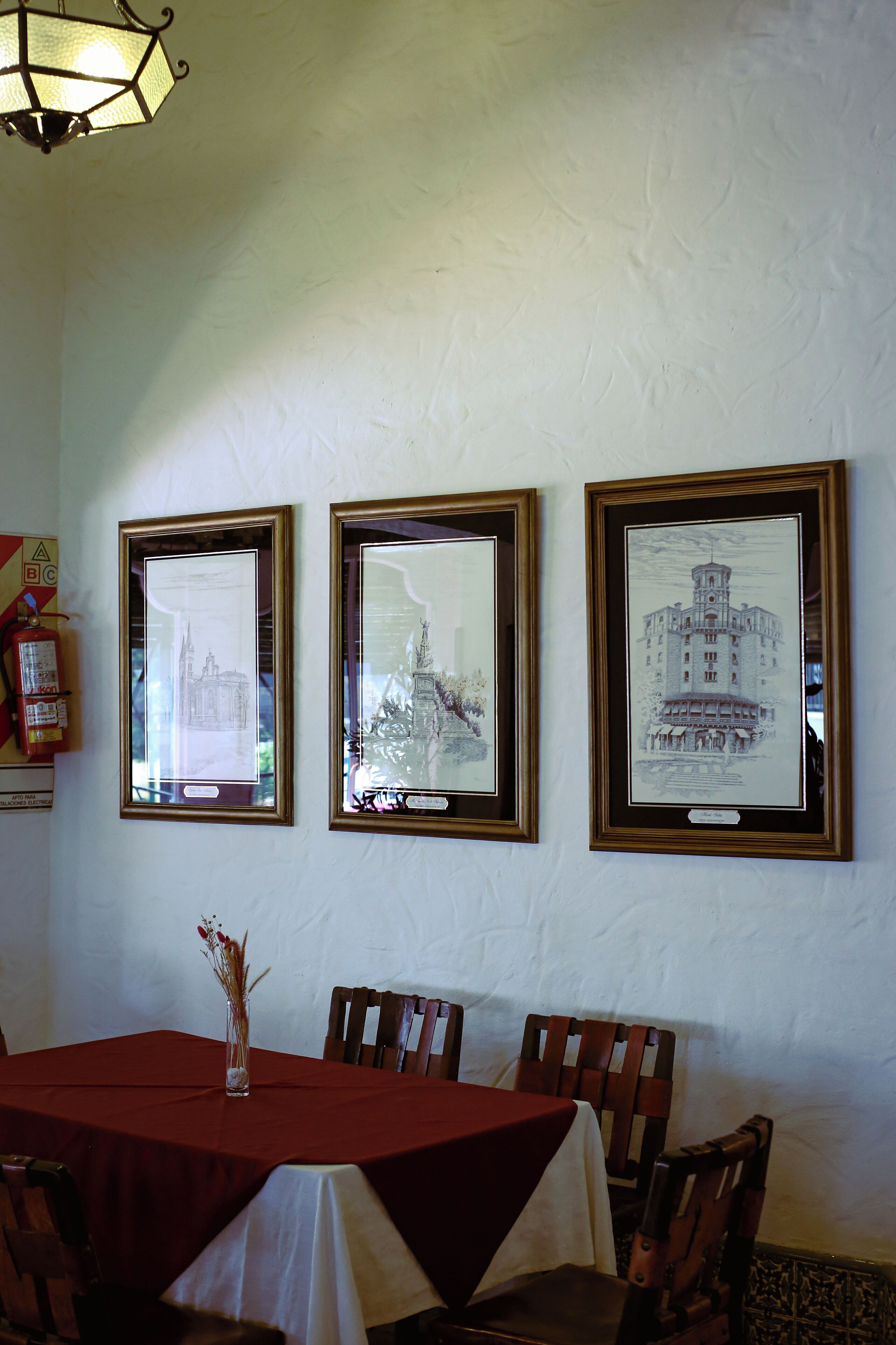 Comedor con una mesa cubierta por un mantel rojo, un cuadro en la pared y un florero con flores secas sobre la mesa.