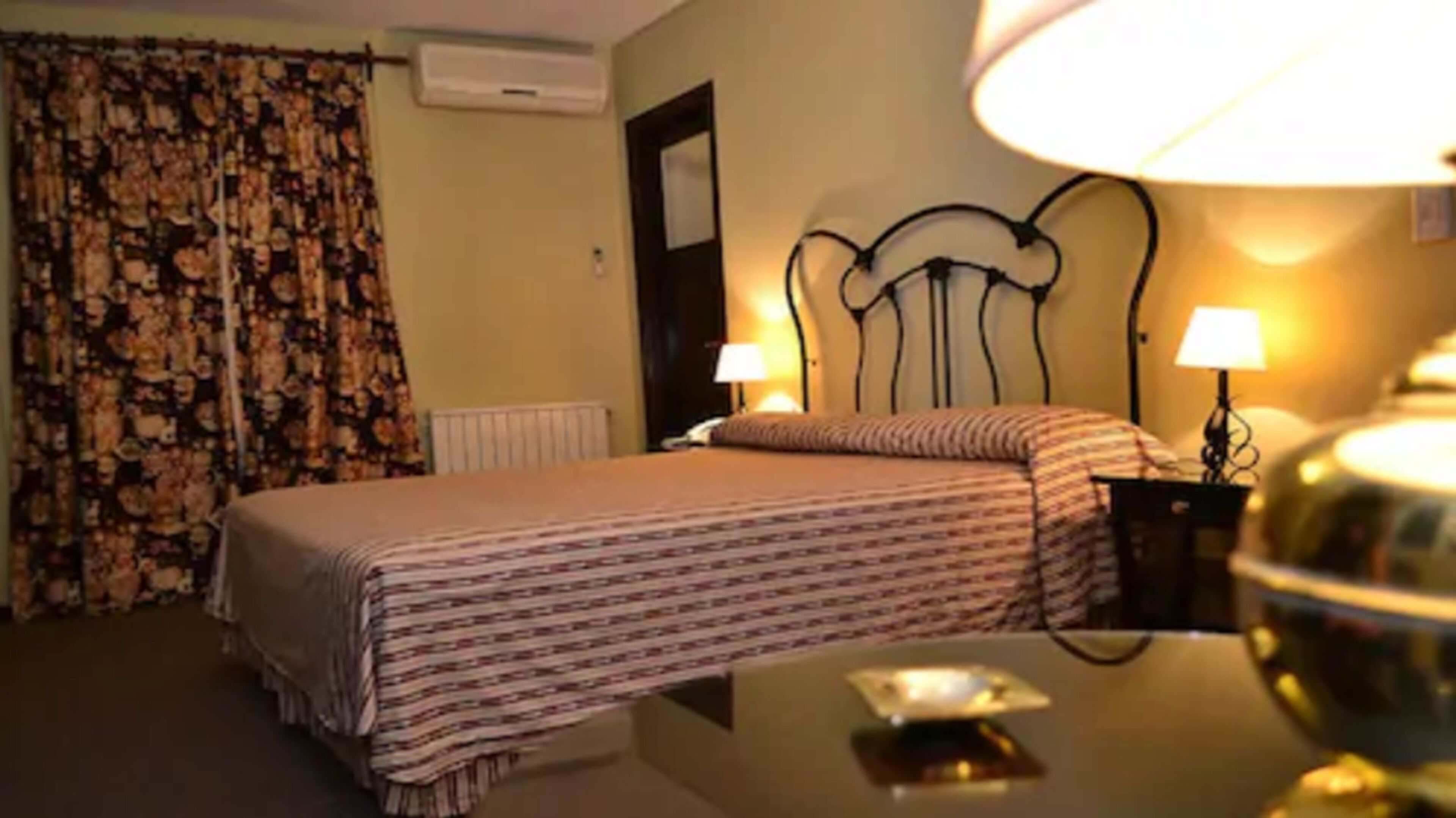 Ein Hotelzimmer mit einem Bett, zwei Nachttischlampen, einem Schreibtisch mit Lampe und einem blumigen Vorhang.