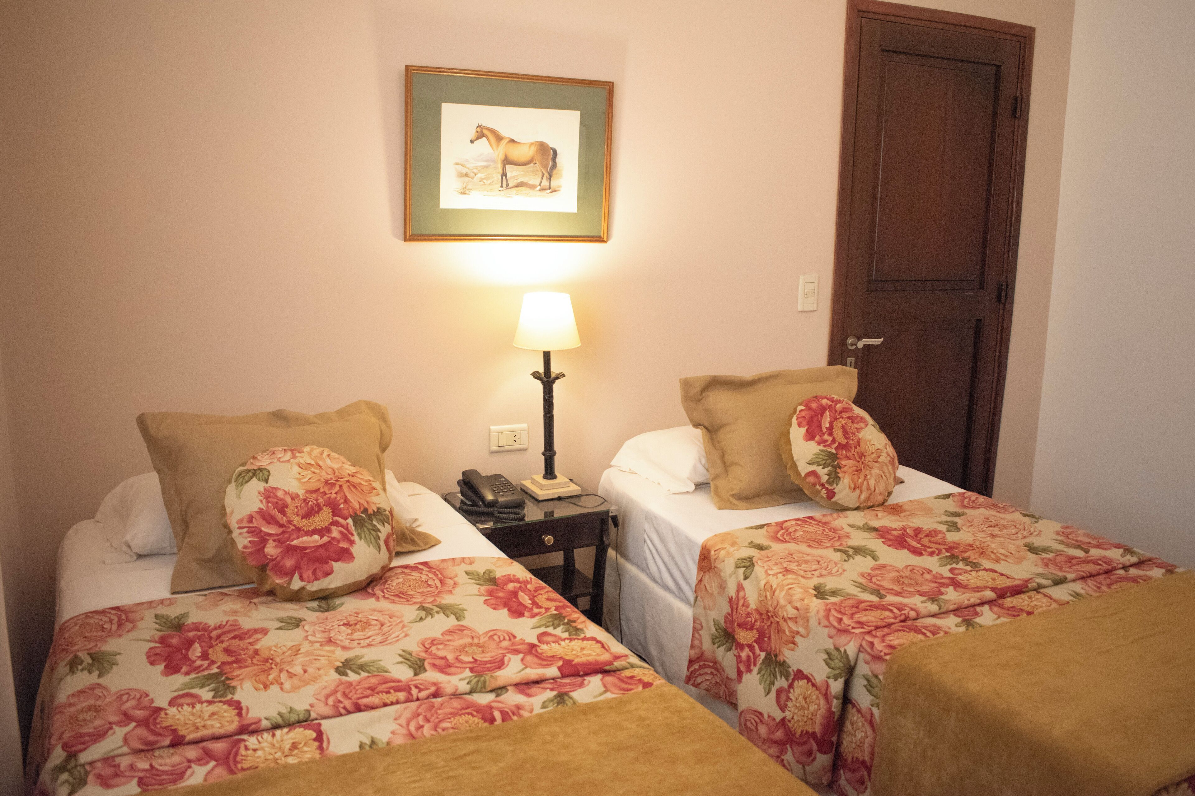 A room with two beds, each with floral bedding and a single pillow. A framed picture hangs on the wall above the beds, and a lamp is on a bedside table.