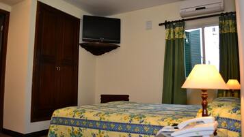 A hotel room with a bed, a desk with a lamp, an air conditioning unit, and a television on the wall.