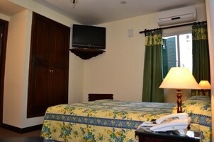 Quarto de hotel com cama, escrivaninha com luminária, ar condicionado e televisão na parede.