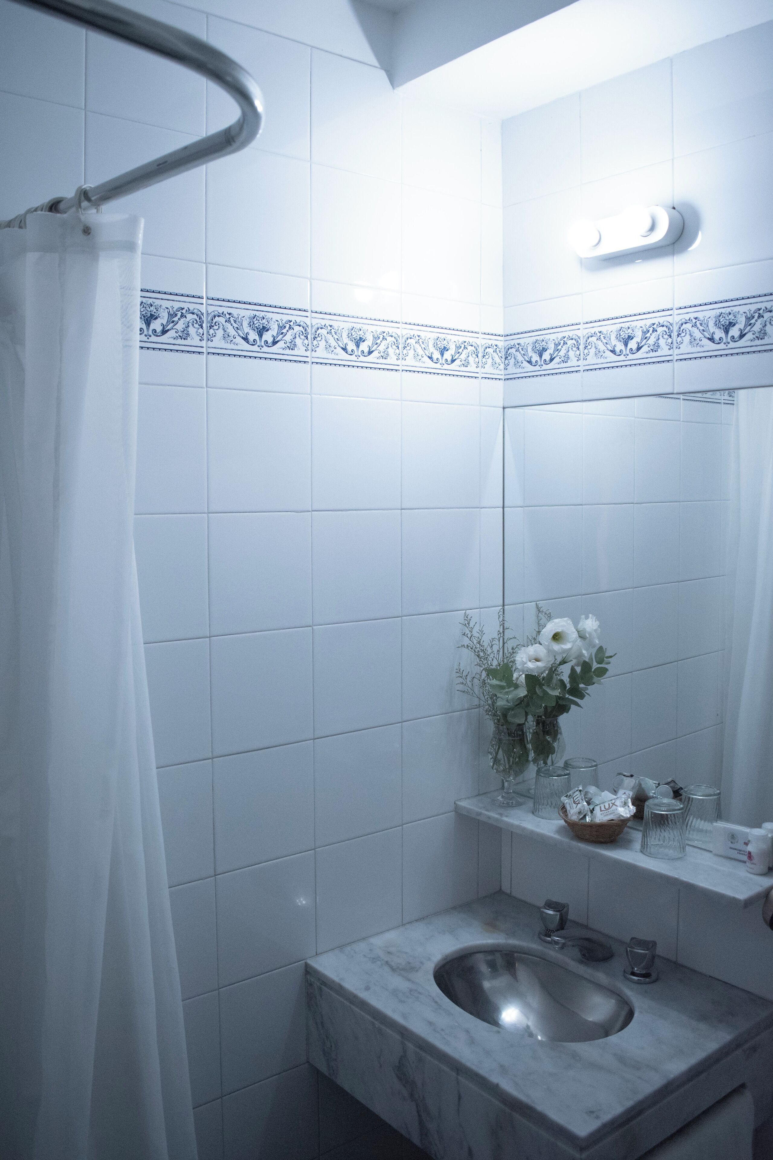 A bathroom with white tiles, a marble sink, a glass vase with flowers, and a shower curtain.