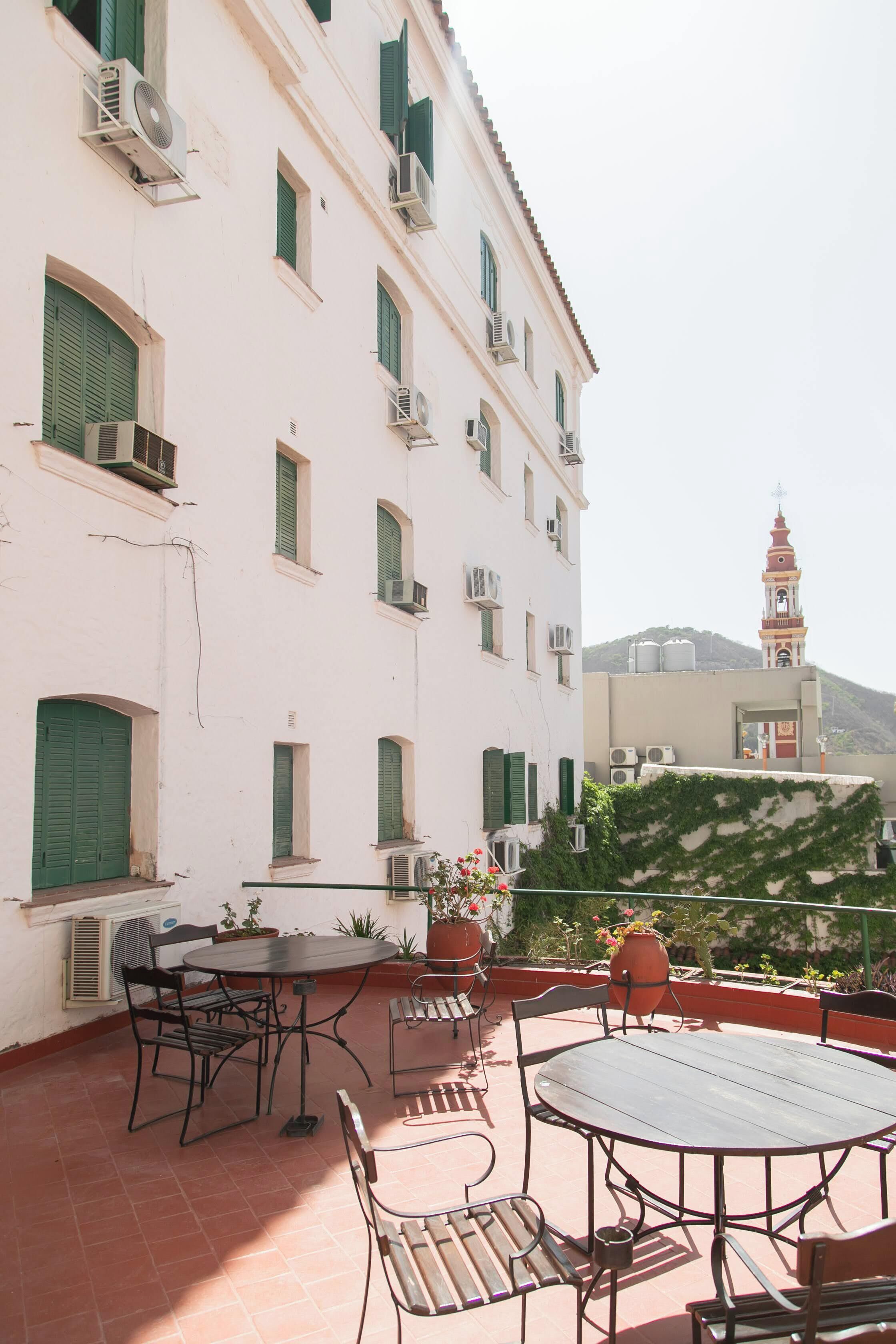 An outdoor patio with a round table and chairs, air conditioning units on the building, and a view of a church tower in the background.