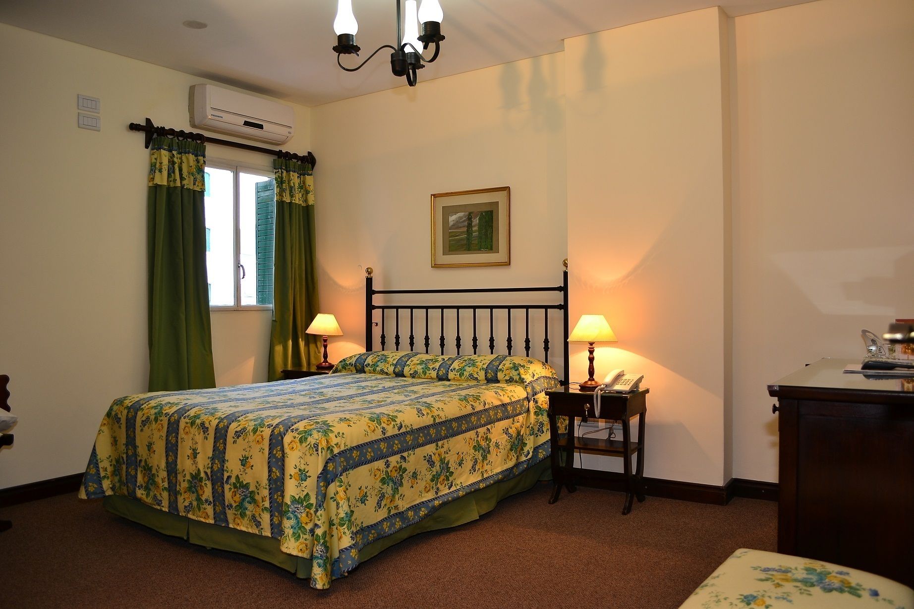 Une chambre à coucher comprenant un lit, deux lampes de chevet, un appareil de climatisation, une fenêtre avec des rideaux et un tableau encadré au mur.