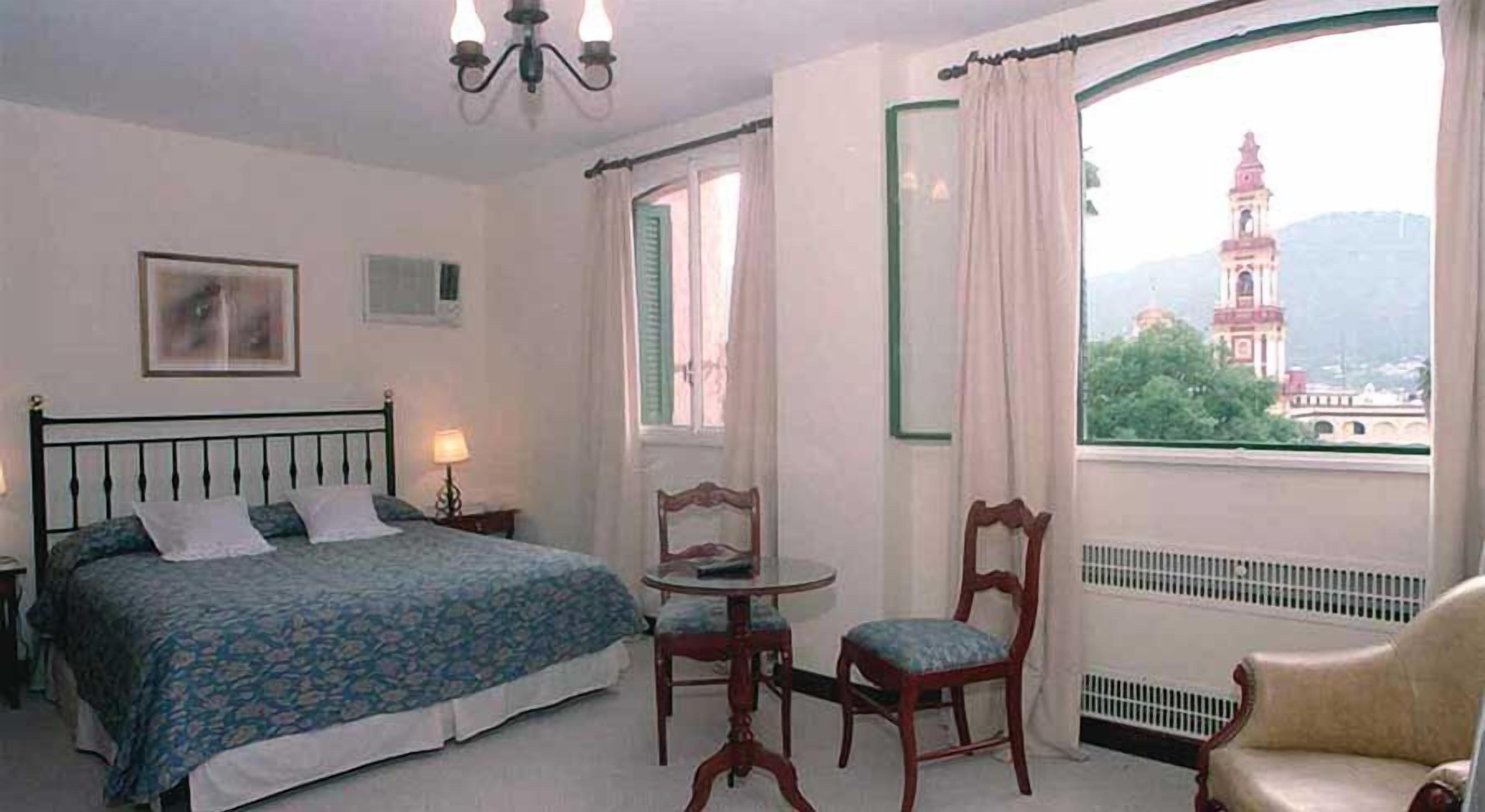 A hotel room with a bed, a chair, a table, and a view of a tower through a window.