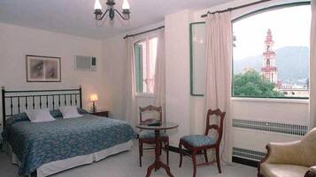 A hotel room with a bed, a chair, a table, and a view of a tower through a window.