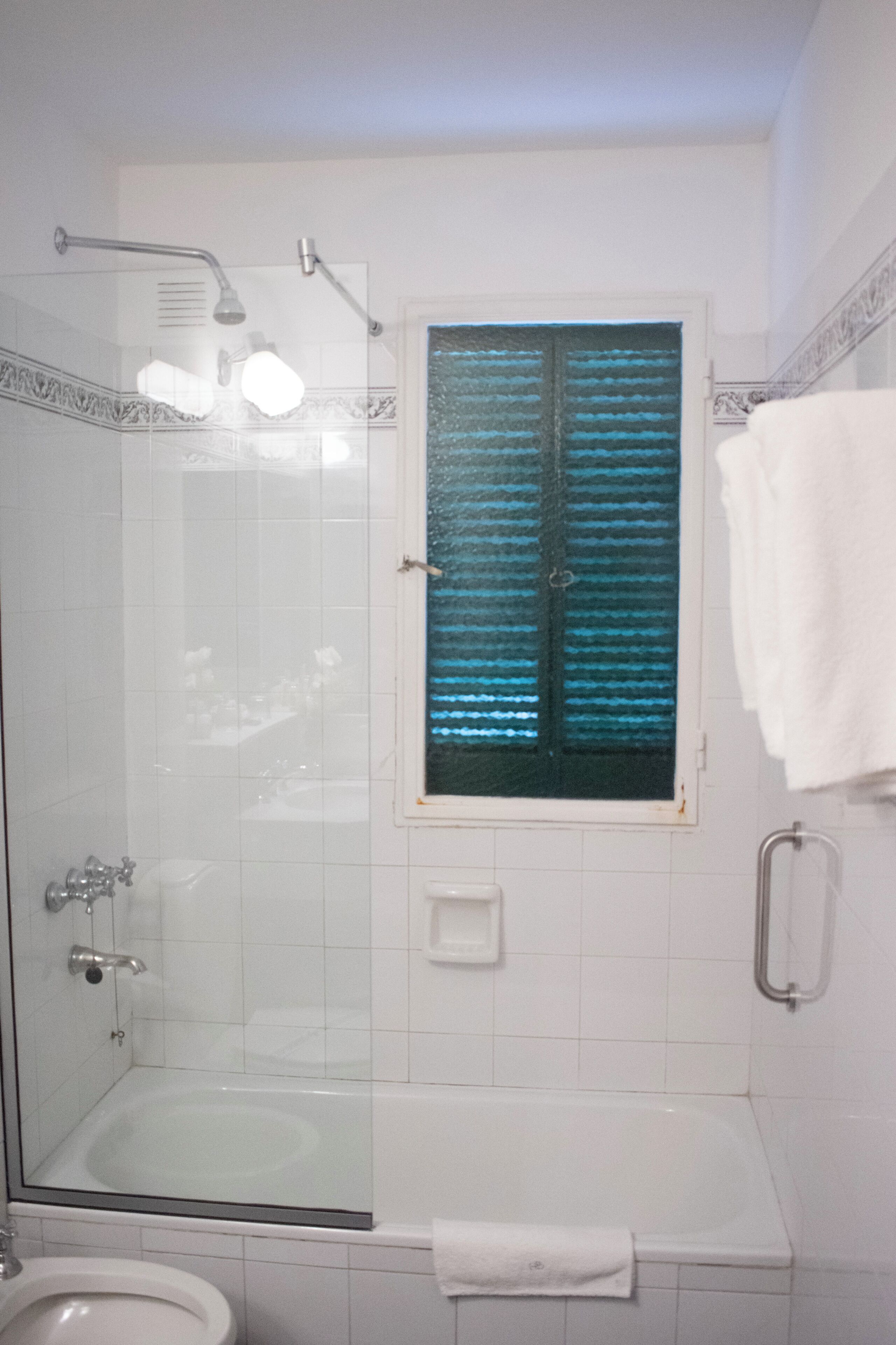 A bathroom with a glass-enclosed shower, a bathtub, a toilet, and a window with blinds.