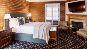 Une chambre d’hôtel avec un mur en briques, un lit avec une tête de lit blanche, deux lampes de chevet, une fenêtre avec des stores, une cheminée, un téléviseur fixé au-dessus de la cheminée et une moquette à motifs.
