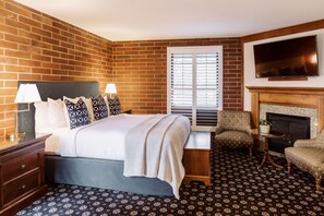 Habitación de hotel con pared de ladrillo, cama con cabecera blanca, dos lámparas de noche, ventana con persianas, chimenea y televisor sobre la chimenea, además de alfombra estampada.