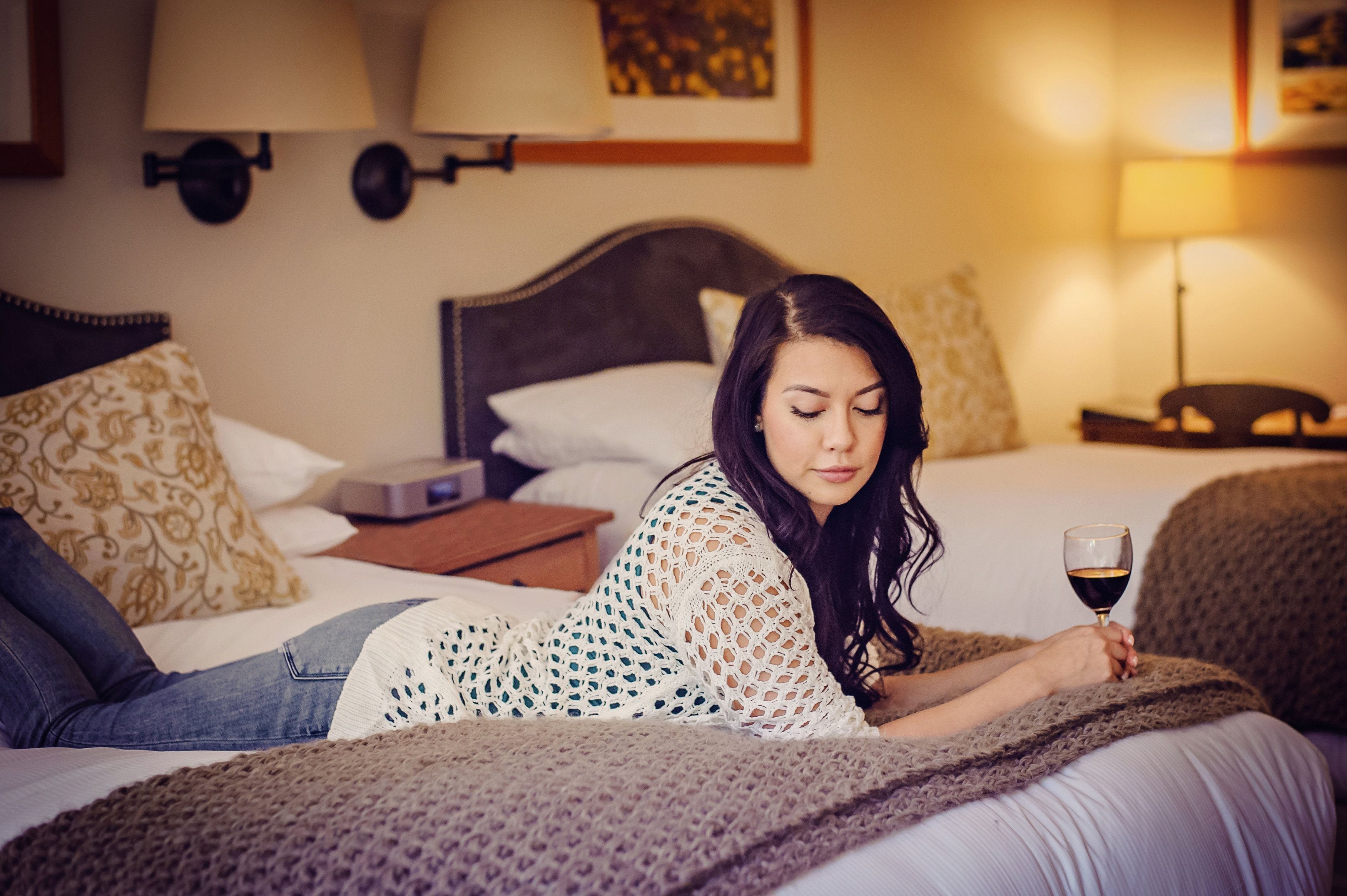 Una persona recostada en una cama con una manta, sosteniendo una copa de vino, en una habitación con cabecera, almohadas y mesita de noche.