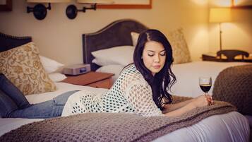 A person lying on a bed with a blanket, holding a glass of wine, in a room with a headboard, pillows, and a nightstand.