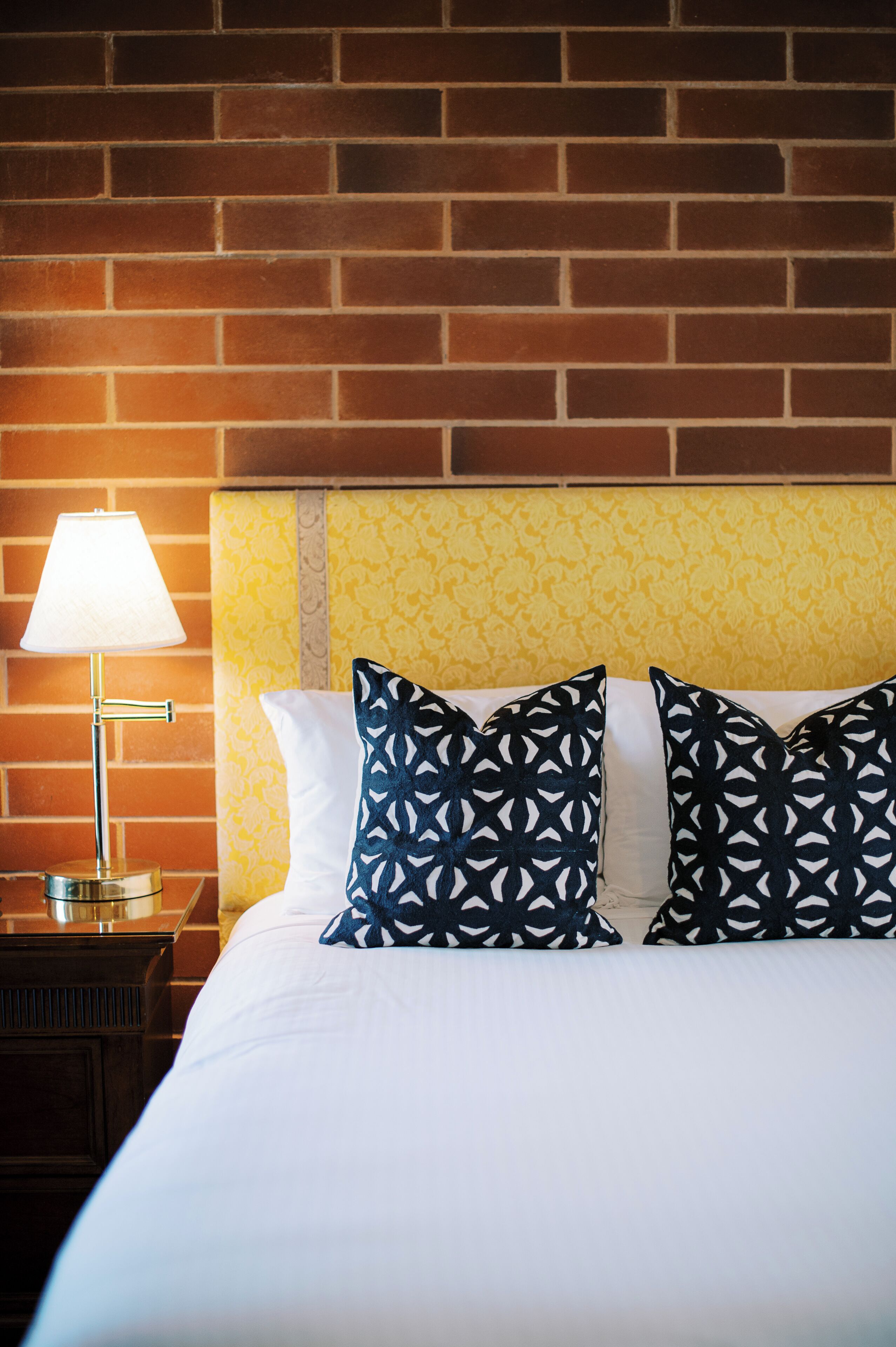 A hotel room with a brick wall, a bed with white bedding, and patterned pillows.