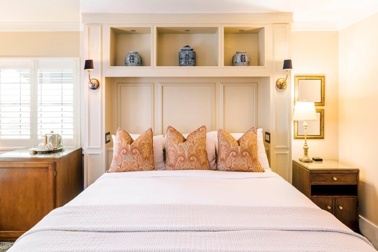 Une chambre d’hôtel avec un grand lit, une table de chevet en bois, une commode, une lampe et des vases décoratifs placés sur des étagères au-dessus du lit.